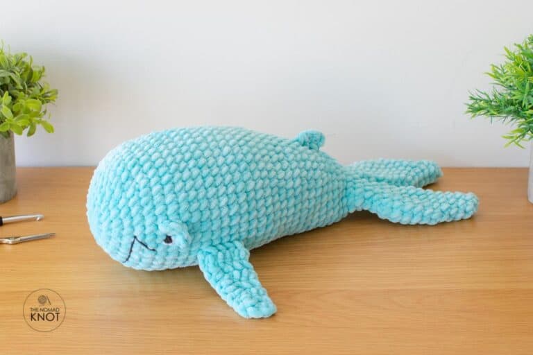 a crochet blue whale inspired free pattern laying on top of a shelf near a crochet hook and scissors.