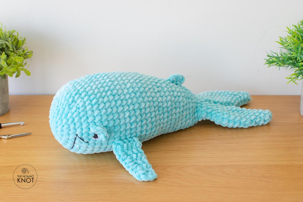 a crochet blue whale inspired free pattern laying on top of a shelf near a crochet hook and scissors.