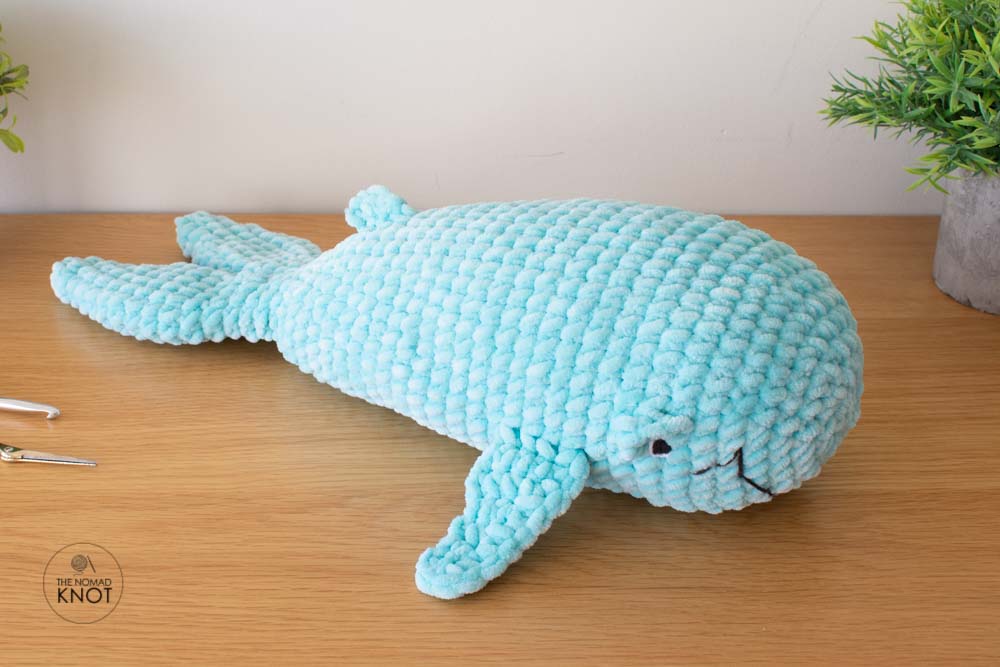 a crochet blue whale inspired free pattern laying on top of a shelf near a crochet hook and scissors.