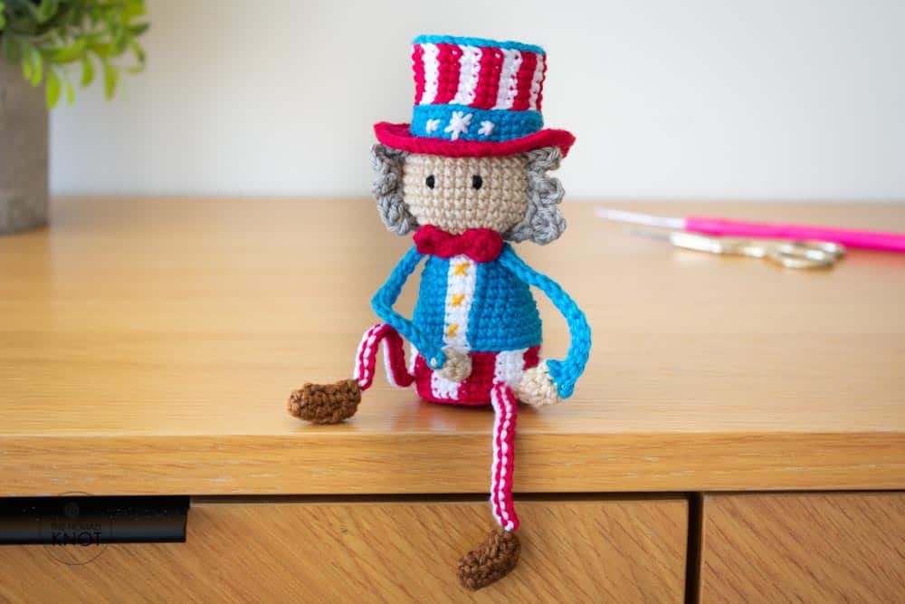 A crochet Uncle Sam free pattern sitting on a shelf with its posable crochet arms on the knee with a plant, scissors and crochet hook on the background