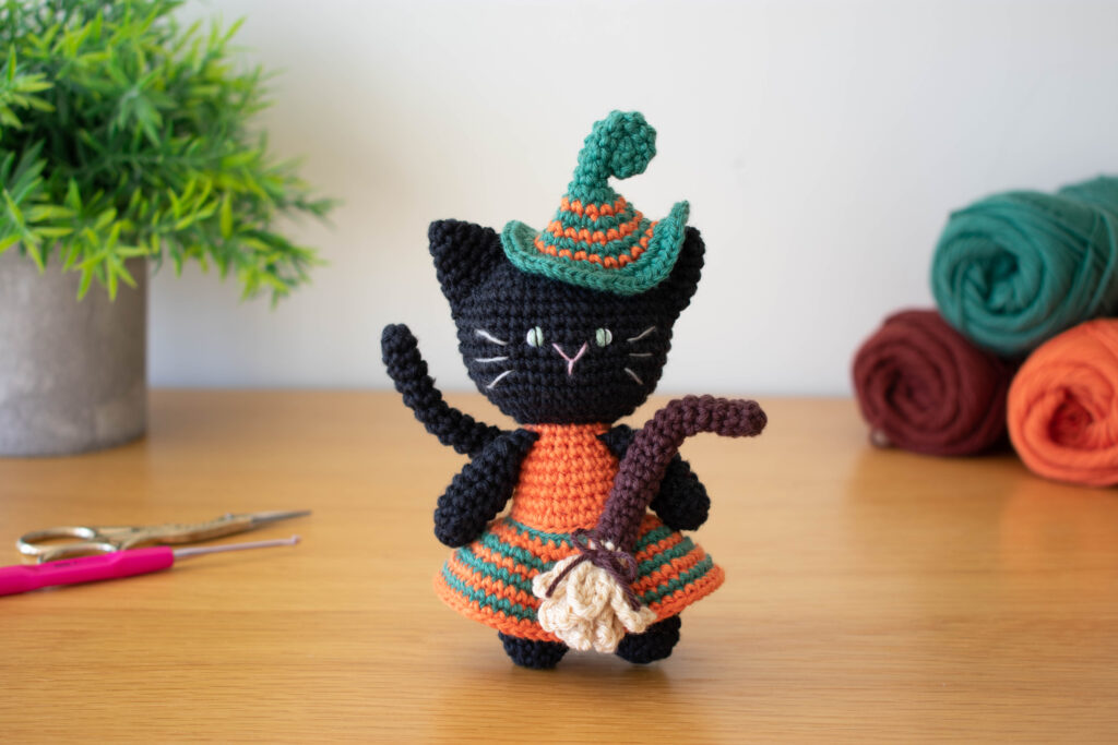 A crochet black cat amigurumi dressed as a witch, featuring a striped orange and green hat and matching dress. The cat holds a small crochet broom and stands on a wooden surface with yarn skeins, scissors, and a crochet hook nearby. A small potted plant adds a cozy touch in the background.