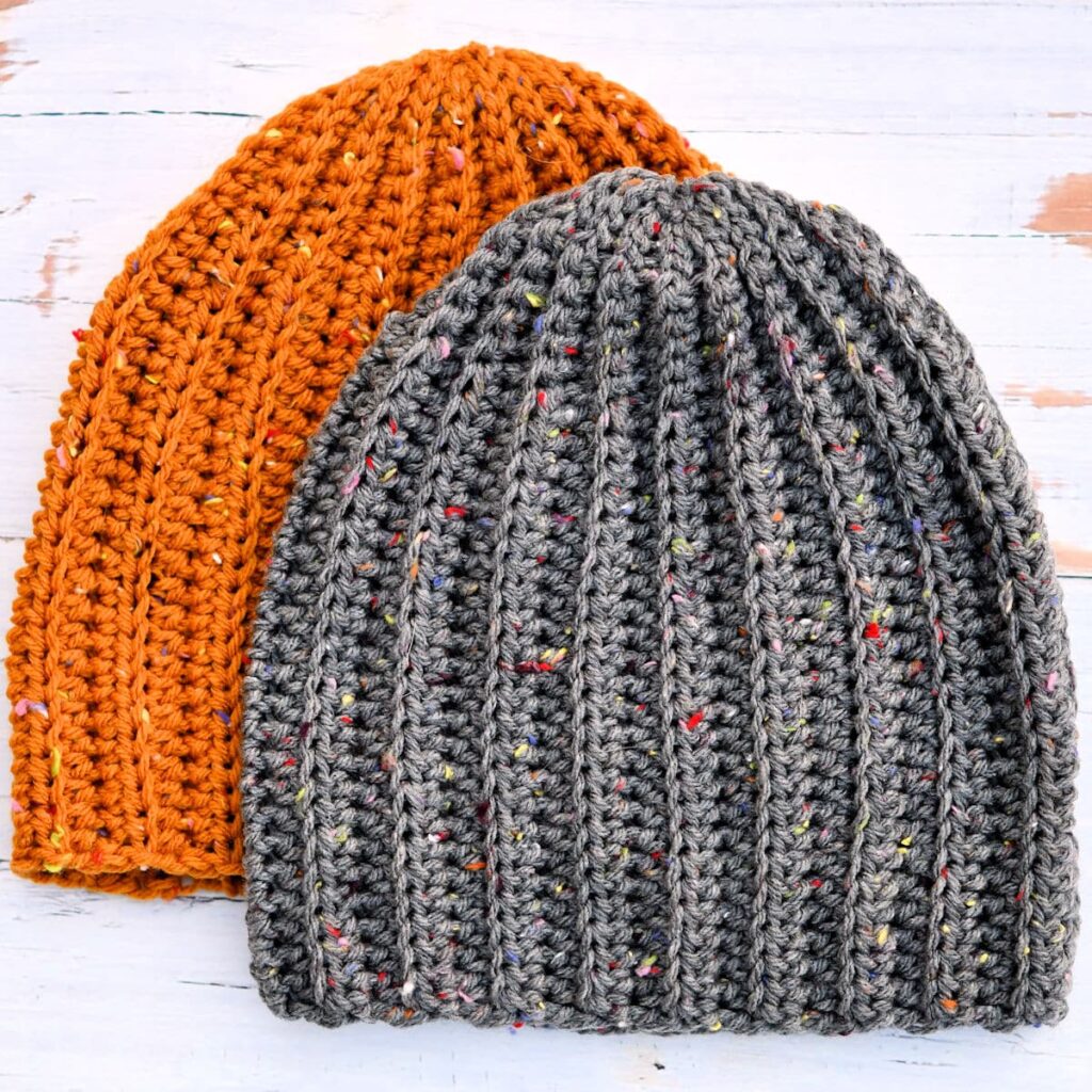 Two ribbed crochet beanies are displayed on a white, distressed wood surface. The beanie in the front is dark gray with colorful flecks, and the beanie partially visible behind it is a flecked, burnt-orange color.