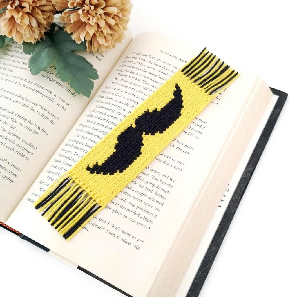 A bright yellow crochet bookmark featuring a large black mustache silhouette in the center lies on the pages of an open book. The bookmark has alternating yellow and black fringe at its end, and a small bouquet of tan flowers rests near the book's spine.