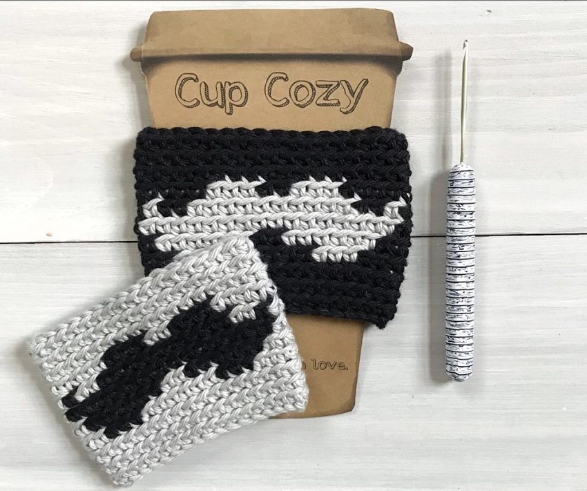 Two crochet cup cozies featuring a buffalo silhouette are displayed on a white wood surface. One cozy is black with a light gray buffalo and is wrapped around a cardboard coffee cup cutout. The other, with the inverse colors of gray with a black buffalo, lies flat nearby. A crochet hook with a patterned handle is placed to the right.