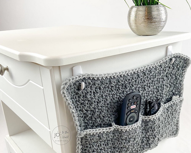 A gray crochet caddy with a light gray border hangs from the side of a white nightstand. The caddy has three pockets, holding a black TV remote in the center pocket and a pair of glasses in the right pocket.