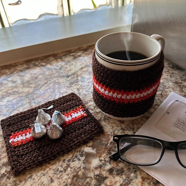 A steaming white mug of coffee, wrapped in a brown crochet cozy with orange and white stripes, sits on a marbled countertop. Beside it is a matching brown rectangular crochet coaster, also with stripes, holding three Hershey's Kisses. A pair of glasses rests nearby.