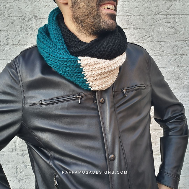 A smiling man wearing a black leather jacket and a thick, ribbed crochet cowl. The cowl is color-blocked with sections of teal, black, and cream, and is pictured against a white brick wall.