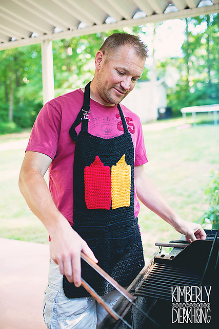 A man wearing a pink t-shirt and a handmade black crochet apron stands at a barbecue grill. The front of the apron is decorated with large, crocheted red ketchup and yellow mustard bottles. He is holding tongs and looking down at the grill with a smile.