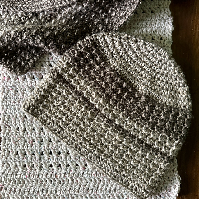 A textured crochet beanie and a matching scarf, both made from variegated brown and gray yarn, are displayed on top of a light gray crochet blanket that has colorful flecks.