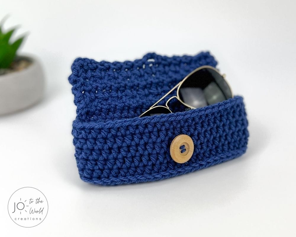 A pair of aviator sunglasses is tucked into a royal blue crochet case. The textured case has a flap closure fastened with a round, light-wood button. The case is sitting on a white surface, with a small potted plant blurred in the background.
