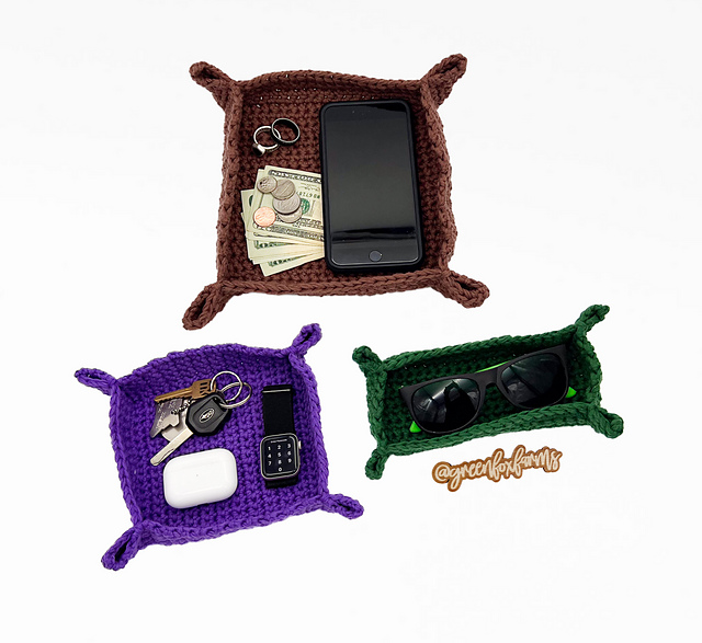 Three square crochet valet trays are displayed on a white background. A large brown tray at the top holds a smartphone, cash, coins, and rings. A purple tray on the bottom left contains keys, a smartwatch, and a wireless earbud case. A smaller dark green tray on the bottom right holds a pair of sunglasses.