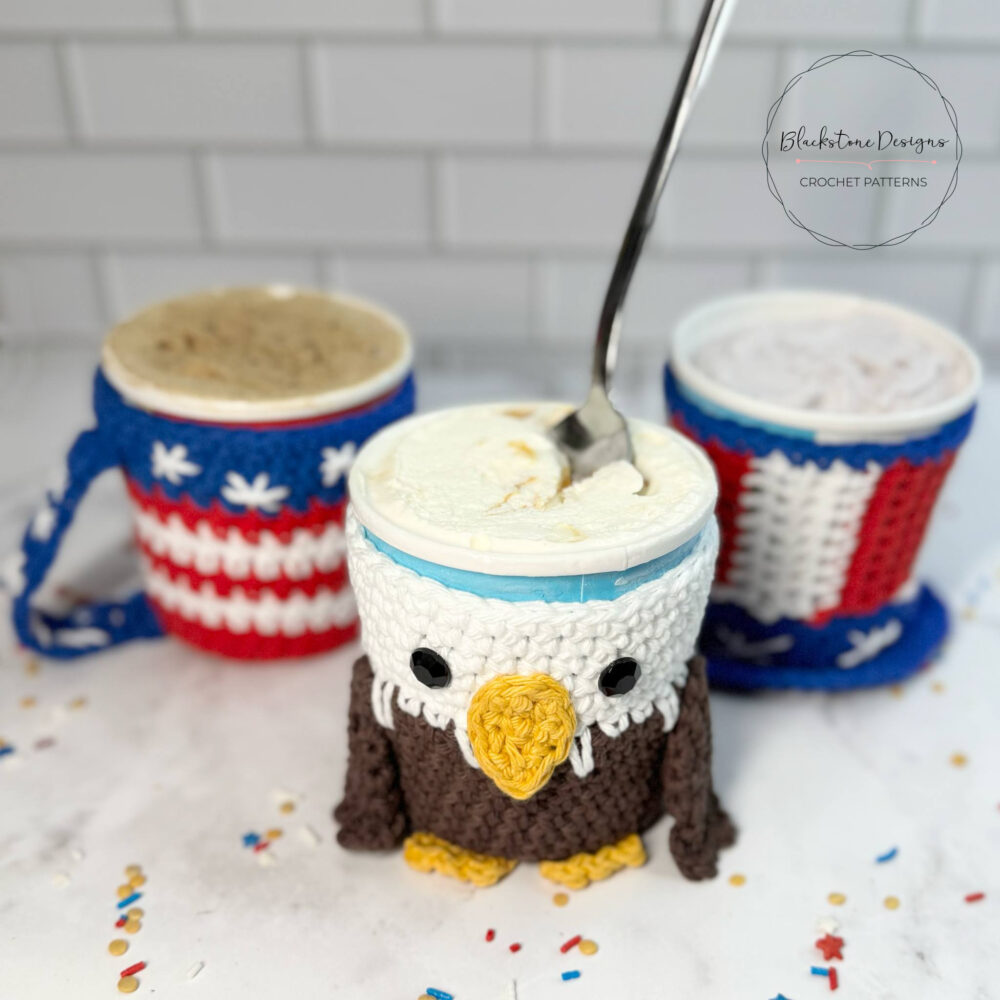A whimsical crochet cozy designed to look like a bald eagle is wrapped around a pint of vanilla ice cream, which has a spoon in it. In the background, two other cozies with American flag patterns are visible: one on a mug and one on another ice cream pint. The items are on a white surface with red, white, and blue sprinkles scattered around.