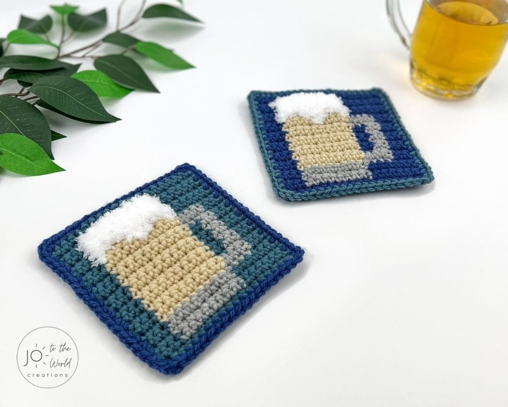 Two square crochet coasters are displayed on a white surface. Each coaster features a tapestry crochet design of a beige beer mug with a gray handle and fluffy white foam, set against a teal and royal blue background. A sprig of green leaves and a glass of beer are visible in the background.