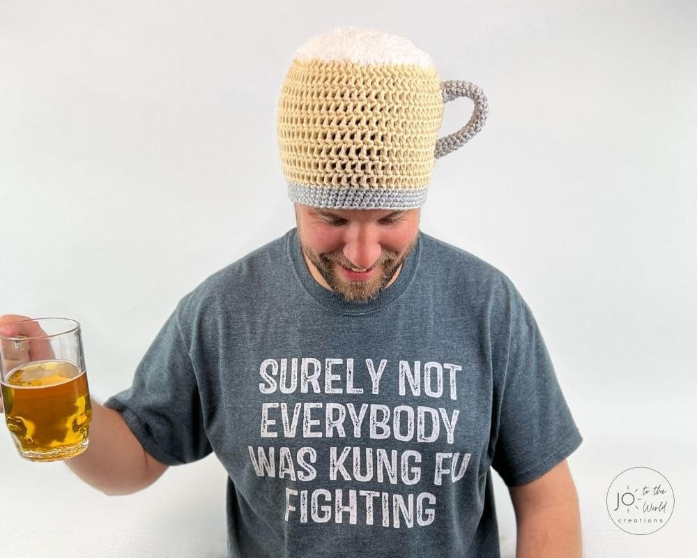 A smiling man wearing a gray t-shirt that reads, "SURELY NOT EVERYBODY WAS KUNG FU FIGHTING." On his head, he wears a novelty crochet hat shaped like a beer mug, complete with light yellow yarn for the beer, fluffy white yarn for the foam, and a gray handle. He is holding a real glass mug of beer in his right hand.