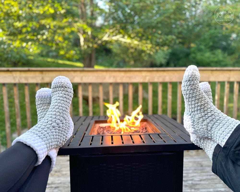 A first-person view of someone's feet propped up in front of a square, black metal fire pit on a wooden deck. The person is wearing a pair of thick, light gray textured crochet socks with fluffy white cuffs. A fire is burning in the pit, and a background of green trees is visible beyond the deck railing.