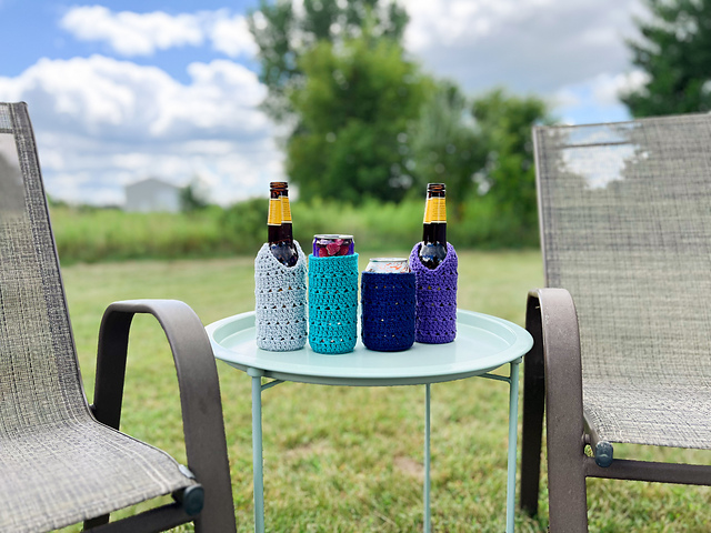Four crochet drink cozies in various shades of blue and purple sit on a light blue metal table outdoors. The cozies are holding two beer bottles and two aluminum cans. The table is positioned on a green lawn between two patio chairs, with a field and trees in the background under a cloudy blue sky.
