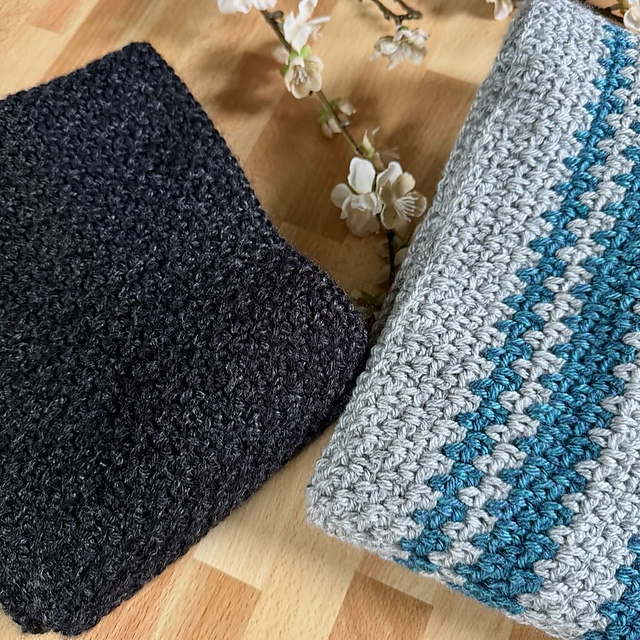 Two men's linen stitch crochet scarves are folded and displayed on a wooden surface. One scarf is solid dark charcoal gray, and the other features stripes of light gray and teal blue. A small sprig of white blossoms rests between them.