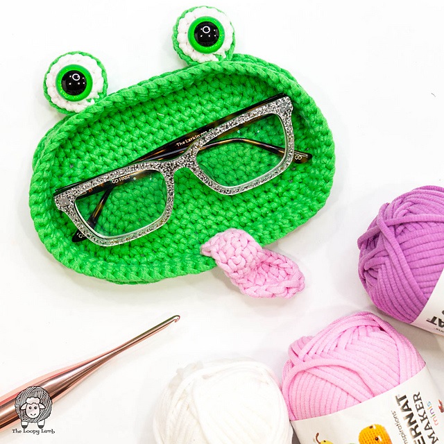 A whimsical, bright green crochet eyeglasses tray shaped like a frog's face, complete with two large, bulging eyes and a small pink tongue. A pair of glittery, tortoiseshell-patterned glasses rests inside the tray. A rose-gold crochet hook and skeins of pink, purple, and white yarn sit nearby on the white surface.