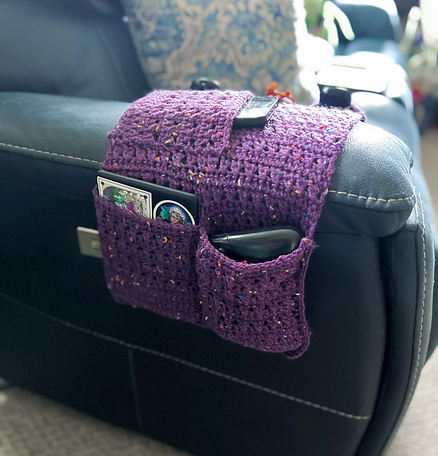 A purple crochet remote caddy, made with yarn that has colorful flecks, is draped over the arm of a black leather recliner. The caddy's multiple pockets are filled with items such as a remote control, a tablet, and a smartphone.