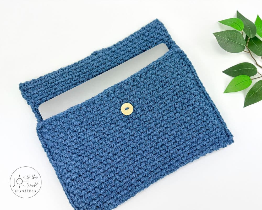 A silver laptop is partially inserted into a textured, blue crochet laptop sleeve. The sleeve has a simple flap closure secured by a small, round wooden button. The items are displayed on a clean white surface, with a sprig of green leaves in the top right corner.